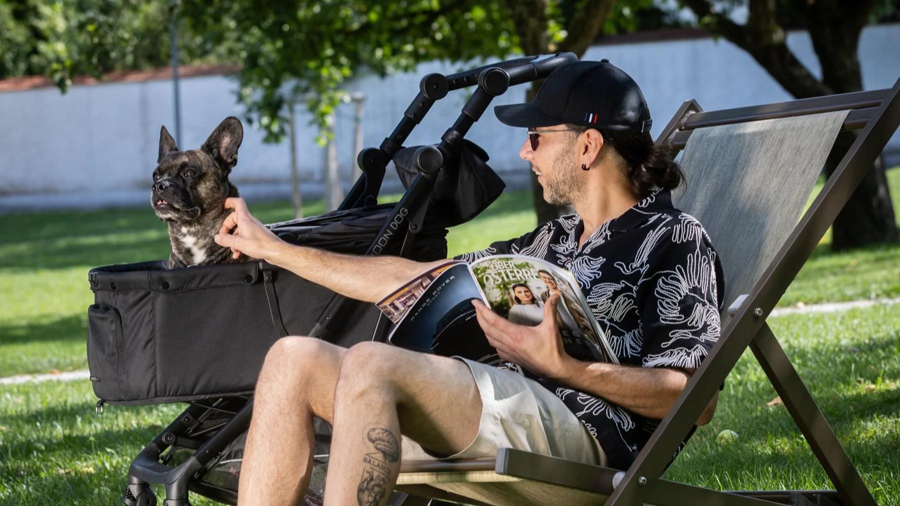 Französische Bulldogge sitzt im MOON Dog Hundebuggy neben ihrem Halter, der im Garten im Liegestuhl liest